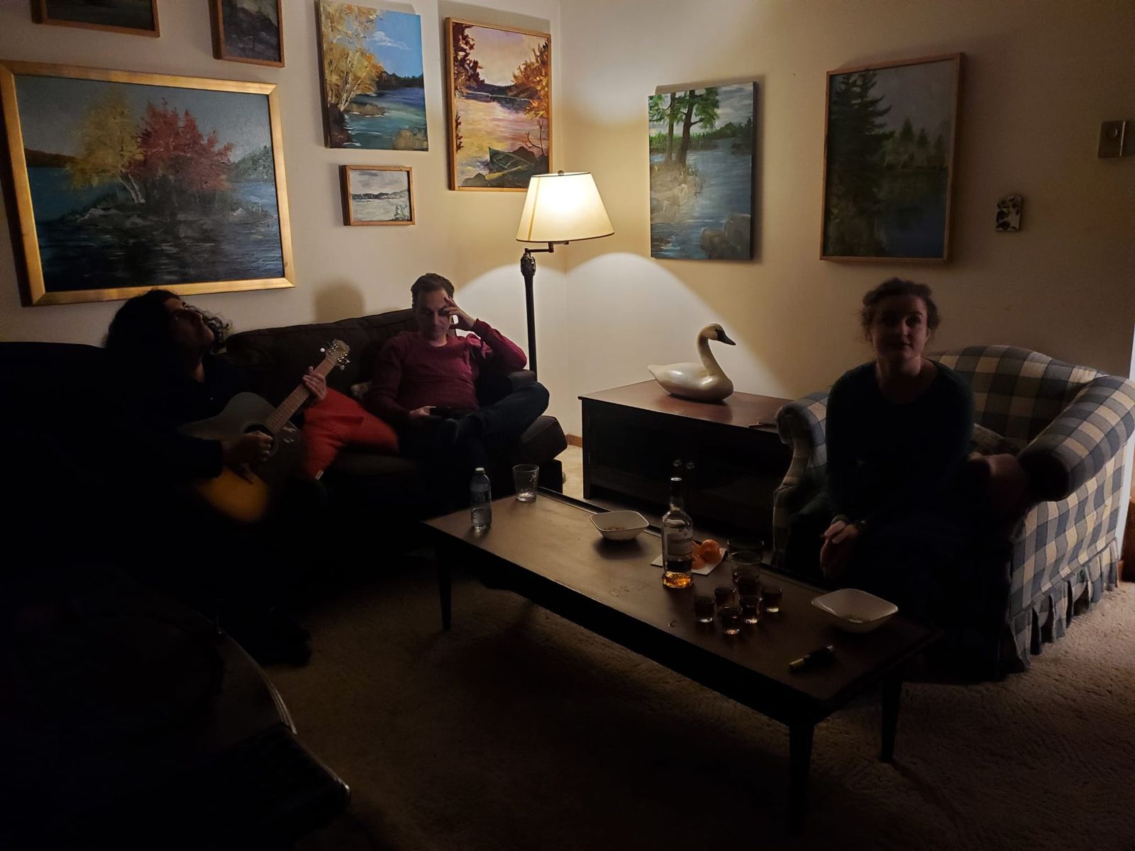 Three cast members in a lamp-lit living room with a guitar