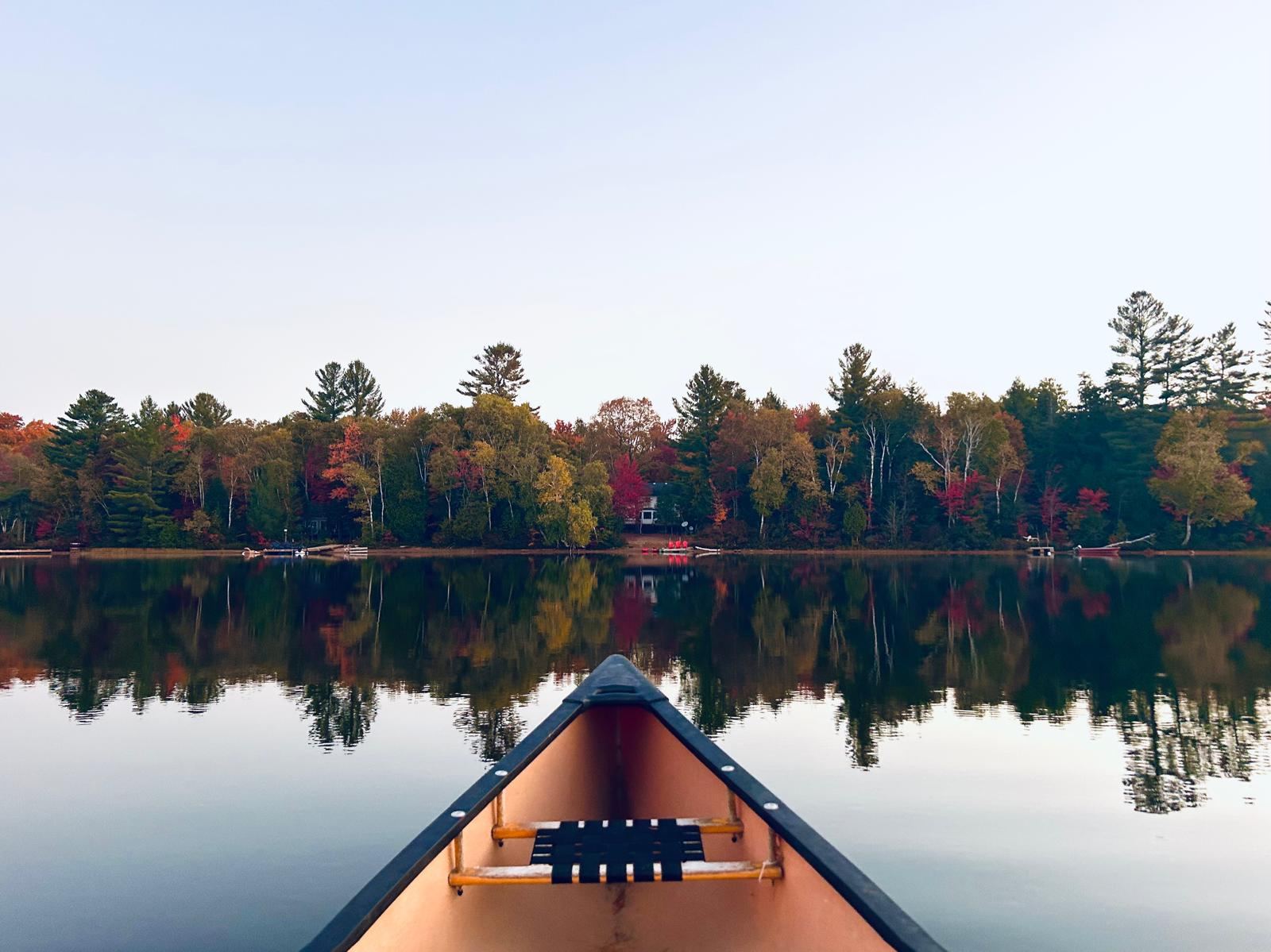 The bow of a canoe pointed at a far shoreline of autumn trees